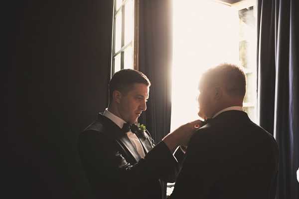 A getting-ready moment captured indoors, showing two men in black tuxedos with white dress shirts near a tall window with bright backlight streaming through. One man, facing the camera, wears a black bow tie and a green boutonniere, and is adjusting or pinning something on the lapel of the other man, whose back is to the camera. Dark navy curtains frame the window, creating strong contrast between the bright backlit window and the dark interior. The composition is a medium portrait shot with dramatic chiaroscuro lighting giving the scene a moody, high-contrast feel.