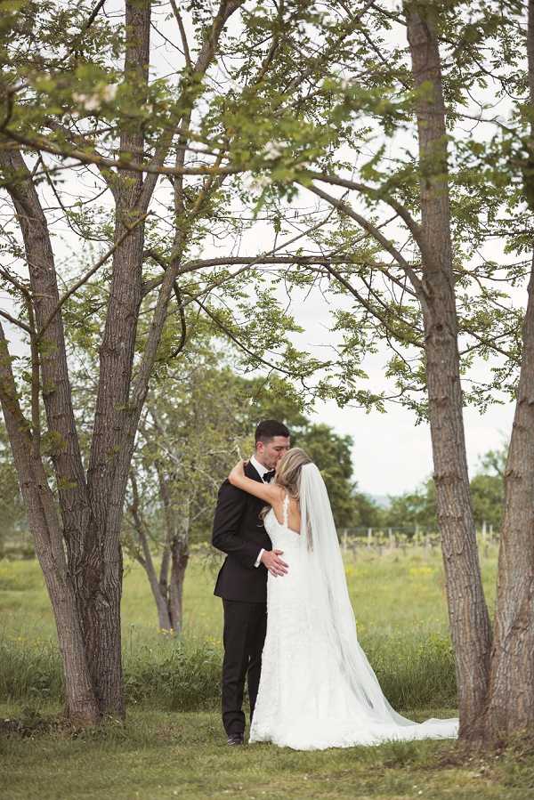 A couple portrait taken outdoors among a grove of trees, with what appears to be vineyard rows visible in the background. The bride and groom are kissing, framed by several tree trunks that create a natural arch around them. The groom wears a black tuxedo with a black bow tie, while the bride wears a fitted ivory lace gown with a low open back and a long cathedral-length veil that trails onto the grass. The composition is a full-length portrait shot with the couple centered between the trees, shot from a slight distance to incorporate the surrounding landscape.