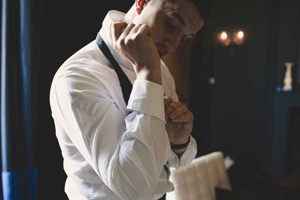 A groom getting ready indoors, fastening his cufflinks on a white dress shirt with dark suspenders visible over his shoulders. He is photographed alone in a dimly lit room with dark walls and warm ambient lighting from a wall sconce visible in the background. The shot is a close-up portrait framing him from roughly the waist up, with moody, low-key lighting creating strong contrast between his white shirt and the dark surroundings. A folded white cloth, likely a pocket square or handkerchief, is partially visible in the lower right corner of the frame.
