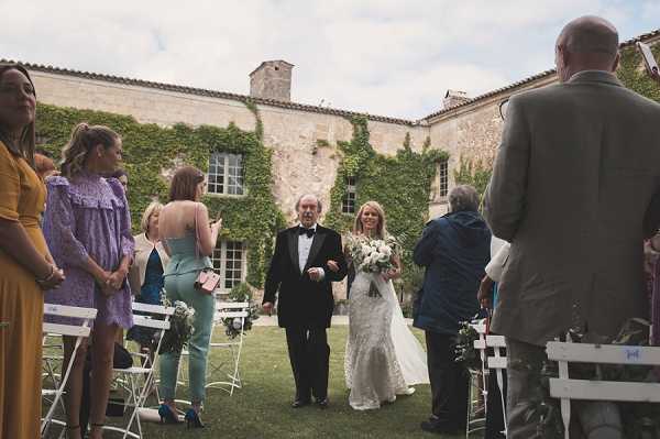 An outdoor wedding ceremony processional taking place in the courtyard of a ivy-covered stone chateau, with the bride walking down the aisle escorted by an older gentleman in a black tuxedo. The bride wears a fitted lace wedding dress and carries a white and greenery bouquet. Guests are seated and standing on either side of the white folding chair-lined aisle, dressed in colorful attire including a mustard yellow dress, a lilac ruffled dress, and a mint green pantsuit. The wide shot captures approximately 15-20 visible guests framing the processional, with the chateau's stone facade covered in climbing ivy serving as the backdrop.