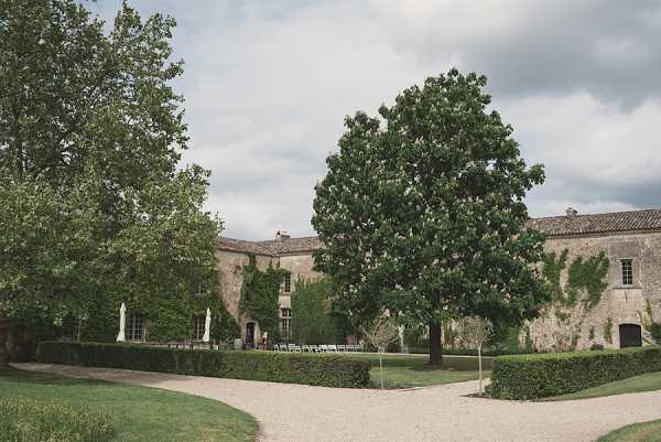 Wide exterior shot of a French stone manor or chateau, likely taken before or between wedding events, with no people currently visible in the main frame. The building features ivy-covered limestone walls, tall shuttered windows, and a terracotta-tiled roof typical of southern French architecture. A row of white folding chairs is set up along the facade, suggesting an outdoor ceremony or cocktail area is being prepared on the gravel courtyard. Potential venue feature image.