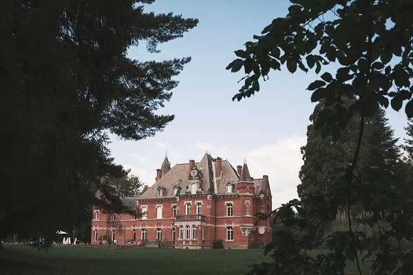Wide exterior shot of a French château venue photographed from across the grounds, framed by large trees in the foreground. The building is a multi-story red brick château with slate-roofed turrets, mansard roofing, and ornate dormer windows typical of 19th-century French architecture. A small white structure or tent is faintly visible to the left side of the building, suggesting event setup. The composition uses the tree canopy as a natural frame around the façade. Potential venue feature image.