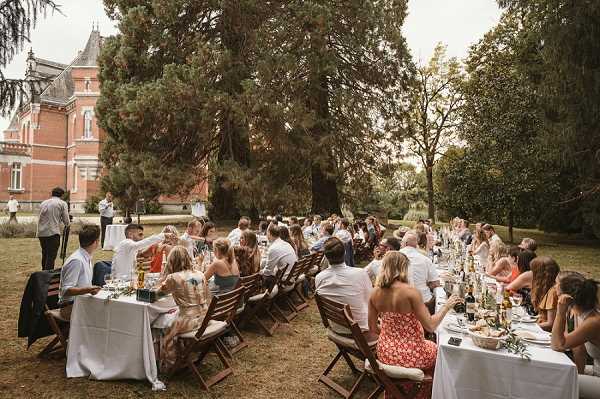 An outdoor wedding reception dinner is underway on the grounds of a red-brick chateau, with approximately 50 guests seated along a single long banquet table covered in white linen. Guests are dressed in summer attire including colorful floral and patterned dresses in red, orange, and yellow tones, with most men in white shirts. The table is set with wine bottles, glassware, and scattered greenery as decor, suggesting a relaxed, garden-party styling approach. The wide shot captures the full length of the table from a slight angle, with the chateau's red-brick facade and tall conifer trees visible in the background. Potential venue feature image.