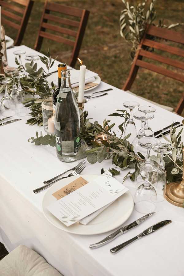 Close-up detail shot of an outdoor wedding reception table set with a white linen tablecloth. The centerpiece features a garland of eucalyptus and olive branches running along the table's length, interspersed with lit taper candles in brass candlestick holders and small votive tea lights. Place settings include white round plates layered with a printed menu card and a kraft paper name card reading 'Lisa,' flanked by silver cutlery and inverted wine glasses. Water and wine bottles are placed along the center of the table, and dark wood folding chairs are visible in the background. The overall styling is minimal and nature-forward with a green and white palette accented by warm brass tones.