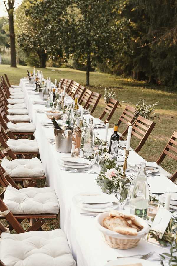 An outdoor wedding reception dinner table set up in a garden or parkland setting, photographed from one end in a perspective wide shot that emphasizes the length of the table. The long rectangular table is covered with a white linen tablecloth and lined on both sides with dark wood folding chairs fitted with white tufted seat cushions. The tablescape features a green garland runner of eucalyptus and olive branches interspersed with blush pink florals, tall white taper candles in brass candlestick holders, silver champagne ice buckets with rosé and white wine bottles, water bottles, wine glasses, white plate place settings, and wicker bread baskets. The decor palette is white, gold, and greenery with blush accents, giving the overall styling a relaxed rustic-chic feel. Fairy lights are faintly visible strung between trees in the background.