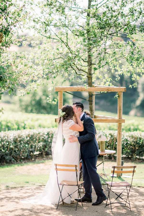 A bride and groom share their first kiss during an outdoor ceremony in a garden setting. The bride wears a white lace cap-sleeve gown with a cathedral veil and has fresh white and green florals woven into her updo, while the groom wears a navy suit. They stand in front of a simple natural wood square arch, flanked by two wooden bistro-style folding chairs and a small round side table. The ceremony setup has a rustic, natural aesthetic with minimal decoration. The shot is a full-length portrait framed by leafy tree branches in the foreground.