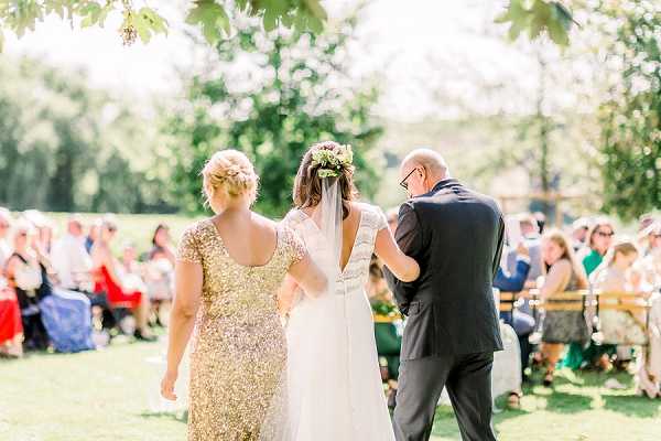 A bride is being escorted down the aisle by two people — a woman in a gold sequined midi dress on her left and a bald man in a dark suit on her right — during an outdoor wedding ceremony. The bride wears a white gown with a low open back, a cathedral-length veil, and a floral crown made of greenery and small blooms. The ceremony is set on a grass lawn with wooden bench seating filled with approximately 30–40 guests dressed in colorful outfits, with a decorated arch or structure visible in the background. The shot is taken from behind the processional group, framed as a medium-wide portrait walking away from the camera.