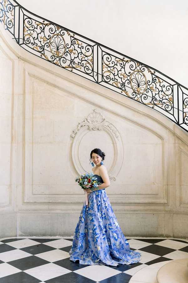 A bridal portrait taken indoors at what appears to be a grand French classical building, featuring a sweeping curved staircase with ornate black and gold wrought-iron railings and carved plaster wall medallion detailing in the background. The bride stands alone on a black and white checkered marble floor, wearing a strapless ball gown in cornflower blue with an all-over floral and botanical print pattern. She holds a loose, garden-style bouquet featuring blue, red, and green mixed flowers, and wears blue drop earrings that coordinate with her dress. Her dark hair is styled in an updo. The composition is a full-length portrait shot, with the diagonal line of the staircase framing her figure against the pale stone wall. Potential venue feature image.