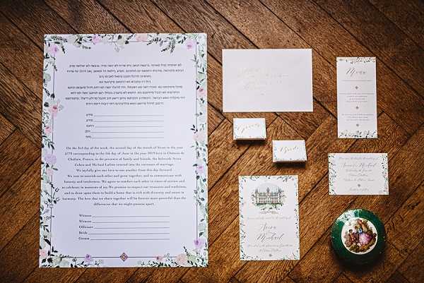 A flat lay close-up shot of a wedding stationery suite arranged on a warm-toned herringbone wood floor. The collection includes a large bilingual ketubah (Jewish marriage contract) with a delicate watercolor floral border in soft lavender, blush pink, and green, a menu card, place cards with gold calligraphy, an invitation suite with matching botanical watercolor detailing, and a save-the-date or information card featuring an illustrated watercolor rendering of a French chateau. A small decorative green trinket box with a painted floral lid is placed in the lower right corner. The overall stationery palette is white and cream with soft floral accents, consistent with a classic yet romantic styling theme.