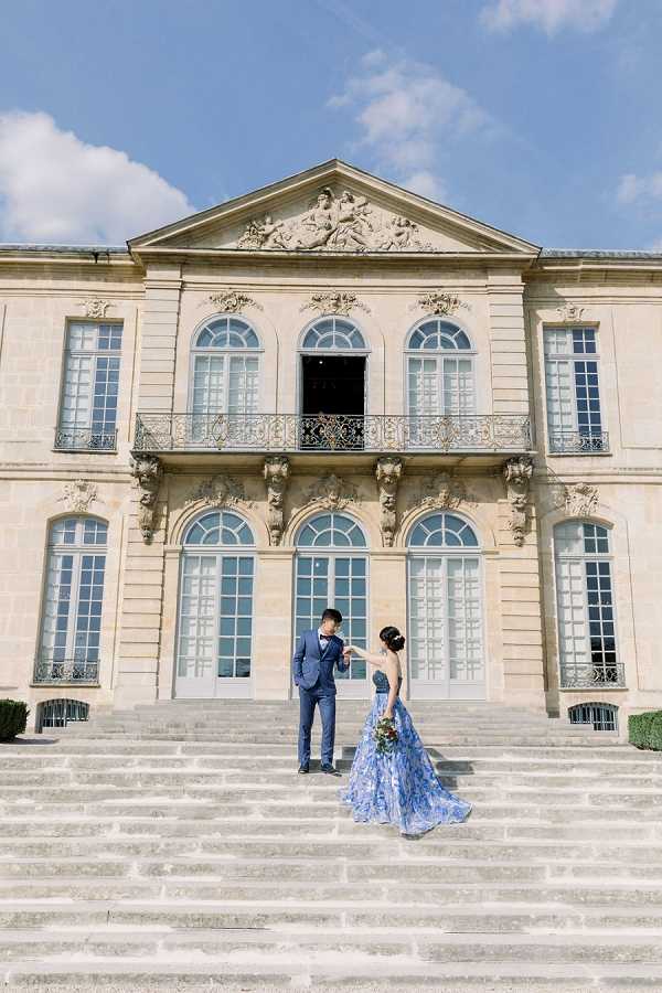A couple poses on the grand stone staircase in front of a classical French neoclassical building featuring arched windows, ornate sculptural pediment, wrought-iron balcony railings, and carved stone detailing. The groom wears a navy blue suit with a bow tie, and the bride wears a floor-length blue floral-print ball gown with a long trailing skirt, holding a small bouquet with warm-toned flowers. The groom holds the bride's hand as she stands a step below him, with the full facade of the building centered behind them. The shot is a wide portrait framing the couple against the full height of the building exterior. Potential venue feature image.