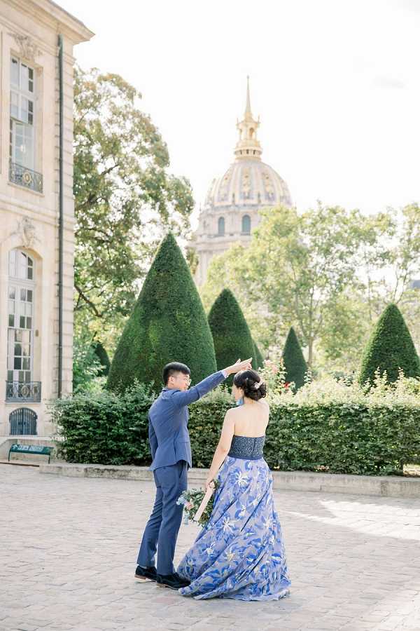 A couple poses outdoors in a Paris courtyard, with the groom spinning the bride as he holds her hand overhead. The groom wears a fitted navy blue suit, while the bride wears a strapless blue floral-embroidered gown with a dark embellished waistband and a flowing skirt; she holds a small bouquet with light pink flowers and greenery and has her hair styled in an updo. The setting features formally clipped conical topiary hedges, a classical French limestone building to the left, and the gold-domed Hôtel des Invalides visible in the background. The shot is a medium-distance portrait taken in natural daylight with a bright, airy exposure.