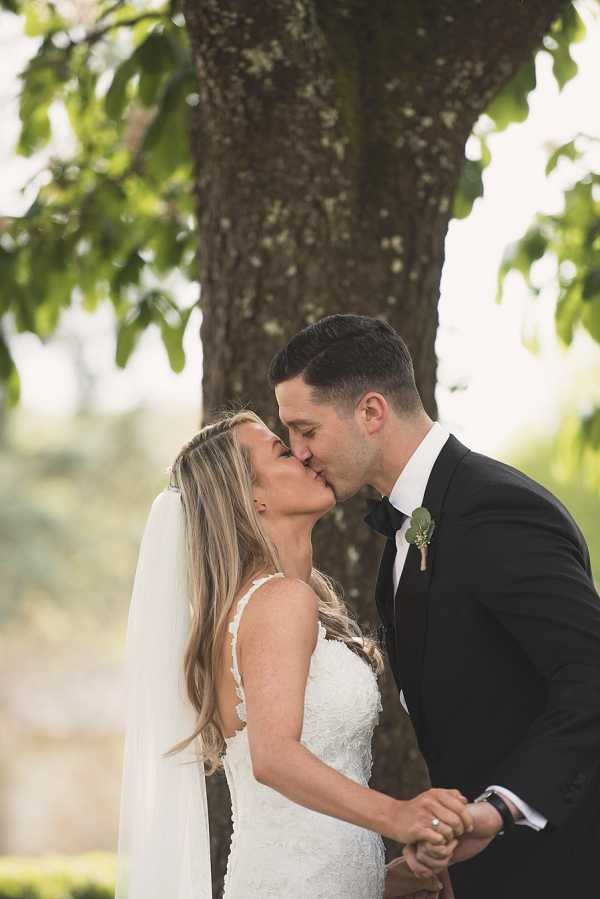 A couple portrait taken outdoors, showing the bride and groom kissing while holding hands in front of a large tree trunk. The bride wears a fitted white lace gown with thin spaghetti straps and an open back detail, with long loose hair and a simple white veil. The groom is dressed in a black tuxedo with a black bow tie and a small greenery boutonniere on his lapel. The shot is a medium portrait framed from the waist up, with a softly blurred green outdoor background.