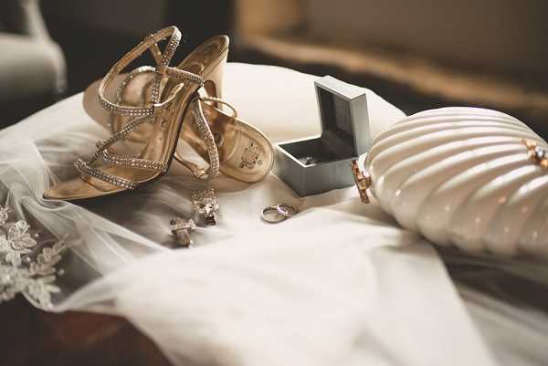 A close-up flat lay of bridal accessories arranged on white fabric, likely the wedding dress or veil. The items include a pair of gold strappy heeled sandals with crystal embellishments, a white shell-shaped clutch bag with gold hardware, a small grey ring box containing wedding bands, a pair of crystal and cross drop earrings, and a lace-edged veil visible at the lower left. The overall accessory palette is gold and white with crystal detailing, consistent with a classic or formal wedding styling.