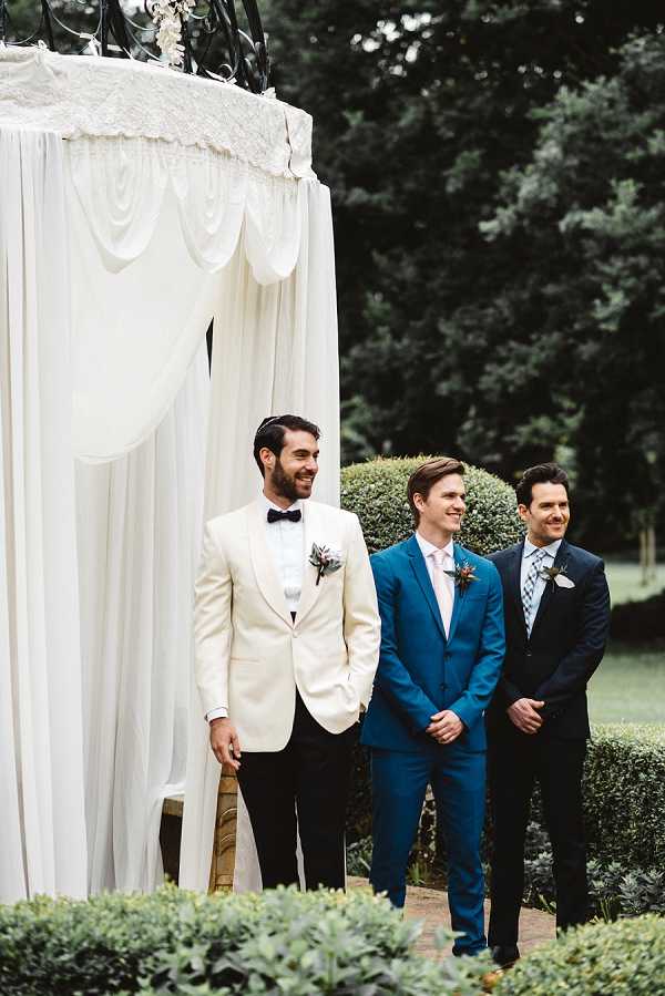 An outdoor ceremony moment showing three men standing at the altar, all wearing boutonnieres. The groom is on the left in a cream/ivory dinner jacket with black bow tie and black trousers, with a small floral boutonniere. The two groomsmen wear a teal/cobalt blue suit with a blush pink tie and a navy suit with a light blue patterned tie, respectively. All three are smiling and appear to be watching the processional. Behind them is a large ceremony structure draped in ivory white fabric with decorative swag detailing and a wrought iron frame visible at the top, with white florals. The setting is an outdoor garden. The shot is a medium portrait framing all three men from roughly mid-thigh up.