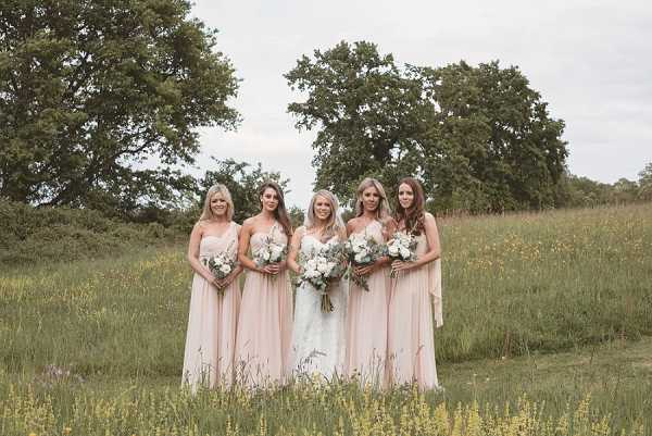 A bridal party portrait taken outdoors in an open meadow, featuring a bride and four bridesmaids standing in a row. The bride wears a strapless white gown and holds a large bouquet of white blooms, while the four bridesmaids wear floor-length strapless blush pink dresses and each carry smaller bouquets of white flowers with greenery. The overall styling is classic and understated, with a soft, cohesive color palette of white and blush pink. The shot is a wide portrait with the group centered in the frame.