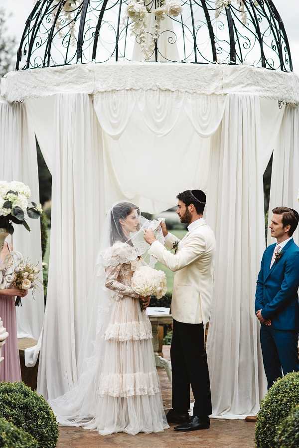 An outdoor Jewish wedding ceremony showing the groom lifting the bride's veil during the bedeken ritual beneath a chuppah. The chuppah is constructed from a black wrought-iron dome-shaped gazebo draped with flowing white fabric panels and topped with white floral arrangements. The bride wears a long-sleeved, tiered ivory and blush gown with lace detailing and carries a full bouquet of white hydrangeas, and the groom wears a cream jacket with black trousers and a kippah. A man in a navy blue suit stands to the right as a witness or officiant, and a bridesmaid in a dusty rose dress is partially visible on the left holding a bouquet of blush and cream blooms. The composition is a medium portrait shot capturing the couple and chuppah in full frame.