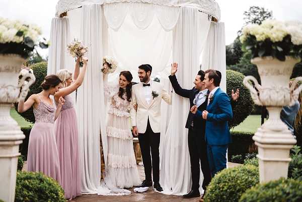 A celebratory moment at the conclusion of an outdoor wedding ceremony, with the couple and their bridal party cheering under a draped white fabric chuppah or ceremony arch adorned with white lace detailing. The bride wears a bohemian-style ivory tiered and ruffled dress with lace accents and holds a bouquet of cream and neutral-toned flowers, while the groom wears a white dinner jacket with black trousers and a black bow tie. Two bridesmaids in dusty rose floor-length gowns stand to the left, and two male attendants — one in a navy blue suit — celebrate to the right, with all members raising their arms in excitement. The outdoor setting features formal garden elements including clipped topiary hedges and ornate white stone urns on pedestals, suggesting a chateau or manor house garden. This is a wide group portrait shot with a documentary feel.