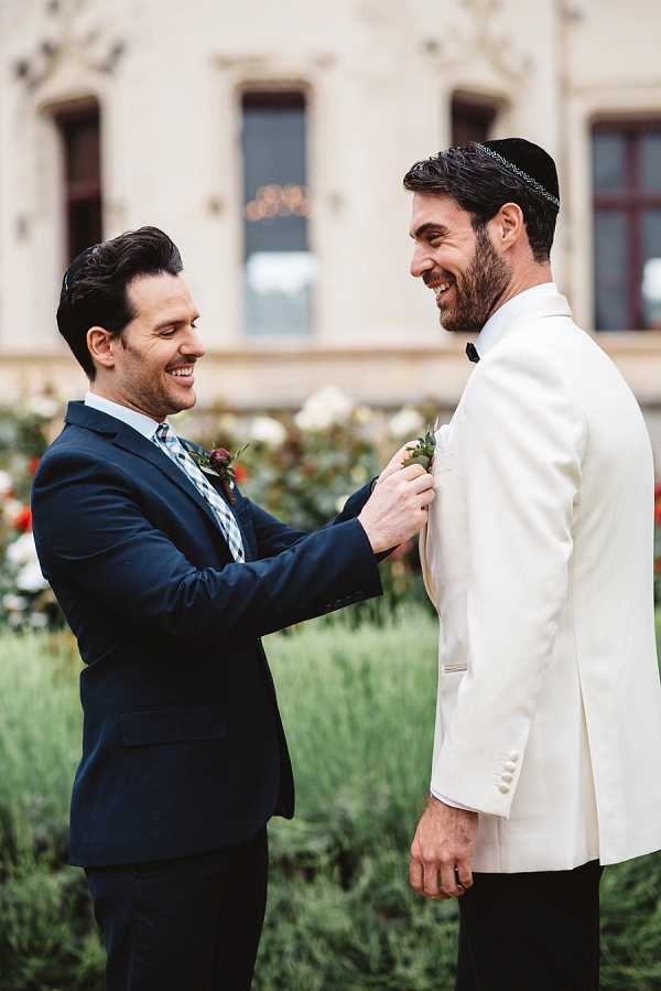A getting-ready or pre-ceremony moment outdoors in the grounds of what appears to be a French chateau, where a man in a navy suit with a patterned tie pins a boutonniere onto the lapel of the groom, who is wearing a white dinner jacket with black trousers and a black kippah. Both men are smiling and laughing during the exchange. The boutonniere features small greenery and what appears to be a deep burgundy bloom. The setting shows garden plantings and the facade of a large period building in the soft-focus background. Portrait shot, medium framing, warm natural light.