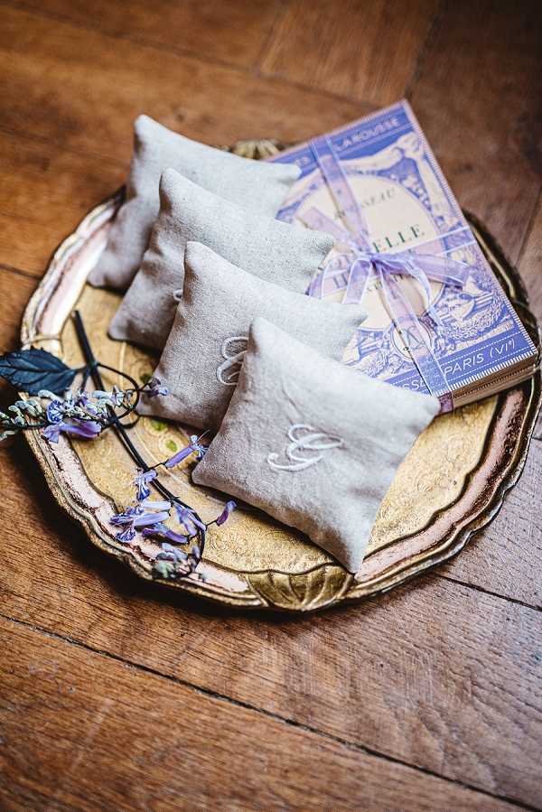 Close-up detail shot of wedding favor items arranged on a gold ornate tray resting on a wooden surface. Four natural linen sachets embroidered with white monogram initials are stacked on the tray alongside a vintage-style Larousse French dictionary tied with a lavender ribbon, referencing a Paris (VIe) address on the cover. A sprig of dried lavender with purple blooms is placed beside the sachets. The overall styling palette is natural linen, gold, and lavender purple, with a French-inspired, rustic-romantic aesthetic.