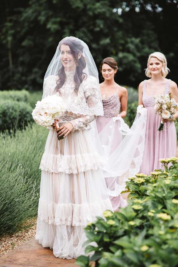 A bride walks along a garden path accompanied by two bridesmaids in an outdoor setting with manicured hedges and lavender in the background. The bride wears a vintage-style ivory tiered tulle gown with lace trim, long lace sleeves, a high neckline, and an embellished belt at the waist, paired with a cathedral-length veil with lace edging draped over her face; she carries a full bouquet of ivory garden roses and peonies. The two bridesmaids wear floor-length dusty mauve dresses — one with a sequined bodice and one with a lace bodice — and carry small bouquets of ivory and blush blooms. The overall styling is romantic and vintage-inspired, captured in a medium portrait shot with soft natural light.