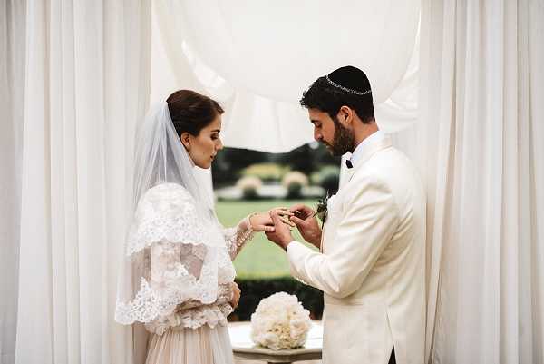 The couple is exchanging rings during their wedding ceremony beneath a draped white fabric chuppah, indicating a Jewish ceremony — the groom wears a kippah. The bride wears a long-sleeve lace wedding gown with a cathedral-length veil, and the groom is dressed in an ivory/cream suit with a black bow tie. A arrangement of white peonies or roses is visible on a small table in the foreground. The ceremony appears to be set outdoors or in an open-air structure, with greenery visible through the fabric draping in the background. The image is a medium portrait shot focused on the ring exchange, with a clean white and ivory color palette throughout the decor and attire.