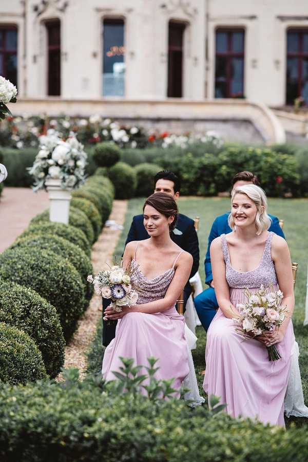 Two bridesmaids are seated during an outdoor ceremony, both wearing floor-length blush pink dresses with silver beaded bodices — one with spaghetti straps and a cowl neckline, the other with a scoop-neck tank style. Each holds a loose, garden-style bouquet featuring white ranunculus, blush roses, and soft greenery with delicate blue accents. Behind them, two male guests in navy and blue suits are visible, seated on gold chiavari chairs along a ceremony aisle lined with neatly trimmed boxwood hedges and white pedestal floral arrangements of white hydrangeas and greenery. The setting is a formal French chateau garden with sculpted topiary and the chateau facade visible in the background. The shot is a medium portrait taken from a low angle along the aisle.