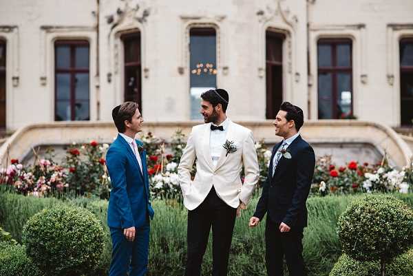 Three men stand in conversation in the formal garden of a French chateau, with the ornate stone facade and grand staircase visible in the background. The groom stands in the center wearing a white dinner jacket, black trousers, black bow tie, a kippah, and a boutonniere with greenery and dark florals. He is flanked by two groomsmen — one in a cobalt blue suit with a pink tie and boutonniere on the left, and one in a navy suit with a blue patterned tie on the right. The garden foreground features neatly trimmed boxwood topiaries and red and white rose bushes. Medium-distance portrait shot with all three men facing each other in relaxed conversation. Potential venue feature image.