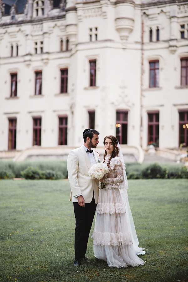 A couple portrait shot outdoors on the lawn in front of a large French chateau with light stone Renaissance-style architecture visible in the background. The bride wears a long-sleeved, tiered ivory tulle wedding dress with lace detailing and holds a rounded bouquet of white peonies and white blooms; the groom wears a cream dinner jacket, black trousers, and a black bow tie, and has a kippah on his head. The two stand close together, facing each other slightly, with the bride looking toward the camera. The composition is a mid-length portrait with the chateau softly blurred in the background, giving depth to the image. Potential venue feature image.