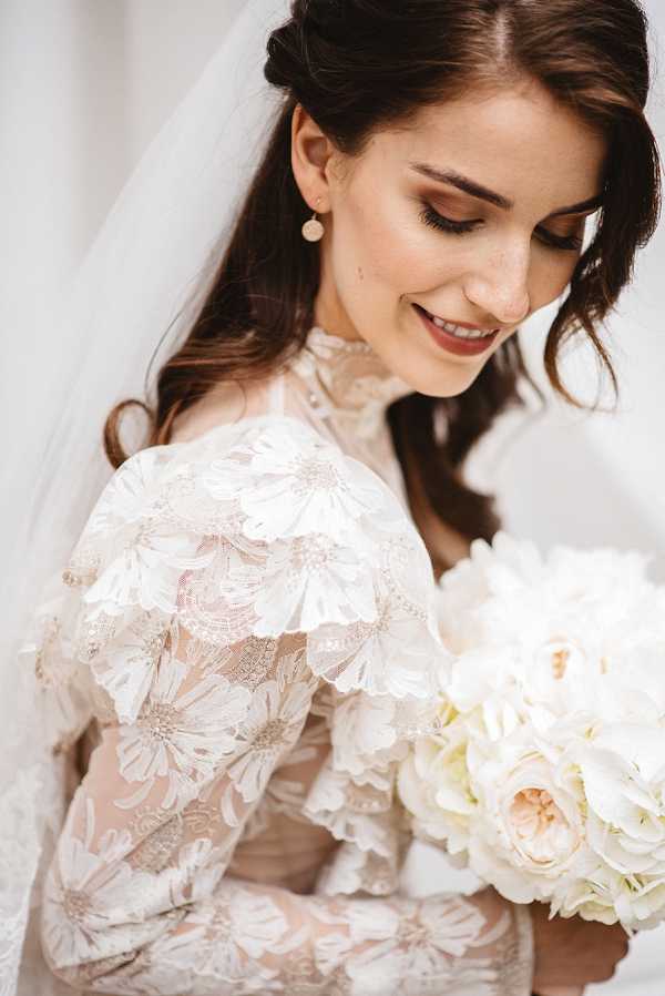 Close-up portrait of a bride looking down and smiling, set against a plain light background. She wears a long-sleeve, high-neck lace gown featuring an appliqué daisy pattern over a nude/blush underlining, giving the dress a floral dimensional texture. Her accessories include small round pearl or stone drop earrings, and she wears a veil visible behind her head. Her dark brown hair is loosely styled half-up with soft waves, and her makeup features warm brown eye shadow and a nude-pink lip. She holds a bouquet of blush garden roses and white hydrangeas with soft cream tones throughout.