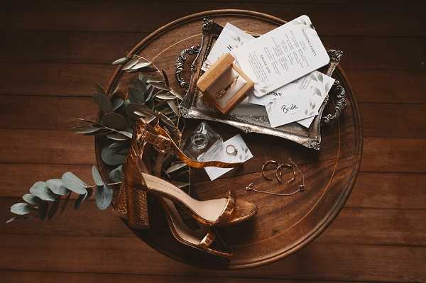 A flat lay detail shot of bridal accessories arranged on a round wooden side table. The items include a pair of gold/bronze metallic platform heels, a silver ornate tray holding a tan ring box, printed stationery cards with botanical-illustrated borders (including a card labeled 'Bride'), a diamond engagement ring displayed on a card, gold hoop earrings, and eucalyptus stems with silver-green leaves. The overall styling palette is warm gold and metallic tones with a rustic, boho aesthetic.