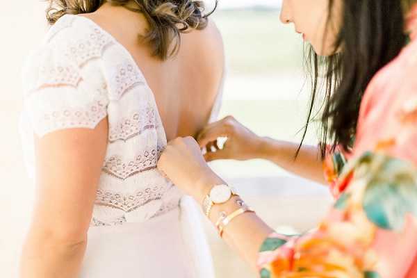 A getting-ready moment captured in a close-up portrait shot, showing a bridesmaid or helper fastening the back of a bride's white lace wedding dress. The dress features a deep open back with lace trim detailing across horizontal panels and short cap sleeves with a bohemian-style aesthetic. The helper is wearing a colorful floral robe in coral, orange, and green tones, along with a rose gold watch and delicate gold and pearl bracelets. The background is bright and overexposed, suggesting an outdoor or well-lit indoor setting.