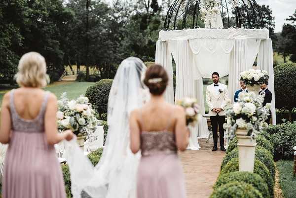An outdoor wedding ceremony captured from behind the bride and two bridesmaids as they face the altar, shot as a wide portrait-style image. The ceremony takes place in a formal garden setting with manicured boxwood hedges lining the aisle and ornamental pedestals holding white floral arrangements. The groom stands at the altar in a white jacket and black trousers, flanked by two attendants, beneath a large black iron gazebo draped with white fabric and topped with a white floral arrangement. The bride wears a cathedral-length veil with her hair pinned up, while the two bridesmaids wear dusty mauve/taupe strapless gowns and carry bouquets of white and cream flowers with greenery. The overall decor palette is white, cream, and soft grey-green, with a classic, formal styling theme.