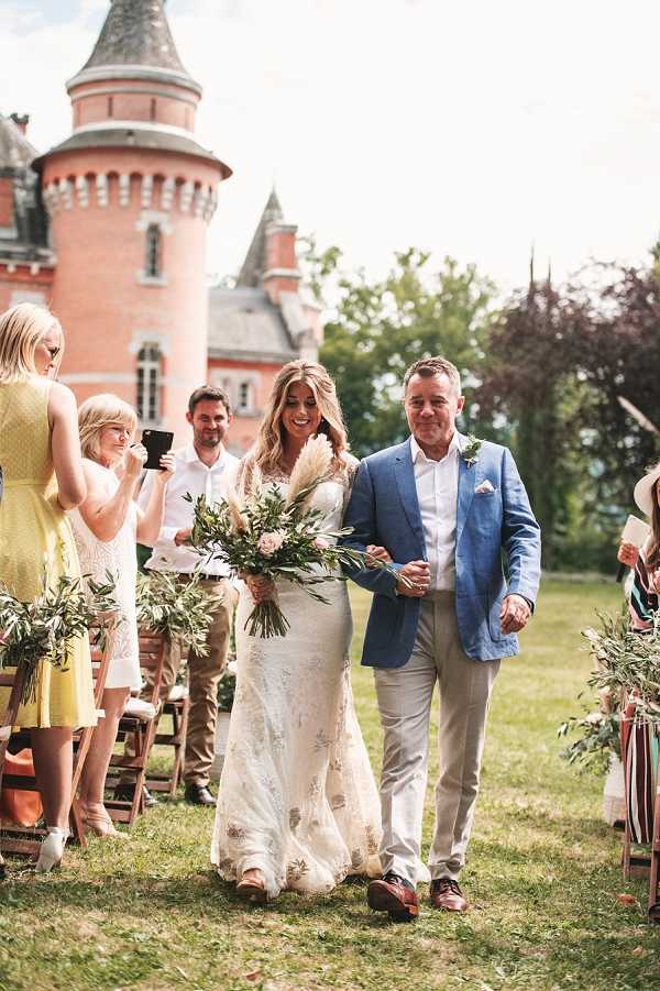 The bride is being walked down the outdoor aisle by a man in a blue blazer and beige trousers, likely her father, during a garden ceremony on the grounds of a red-brick French chateau with a distinctive round turret visible in the background. The bride wears a fitted, long-sleeved lace gown with floral embroidery and carries a large, loose bouquet featuring pampas grass, greenery, olive branches, and blush blooms in a boho style. Guests seated on wooden folding chairs line both sides of the grass aisle, with olive branch arrangements decorating the chair ends; onlookers including women in yellow dresses are visible to the left. The shot is a medium wide portrait taken at aisle level, capturing the processional moment with the chateau architecture prominently featured in the background. Potential venue feature image.