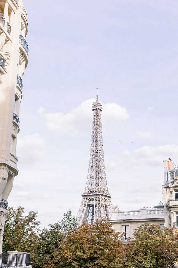 A wide-angle outdoor location shot in Paris featuring the Eiffel Tower rising between classic Haussmann-style cream stone buildings with wrought iron balconies. No people or wedding subjects are visible in the frame. The composition is a vertical wide shot captured from street level, with the iron tower centered between the surrounding architecture. Potential venue feature image.