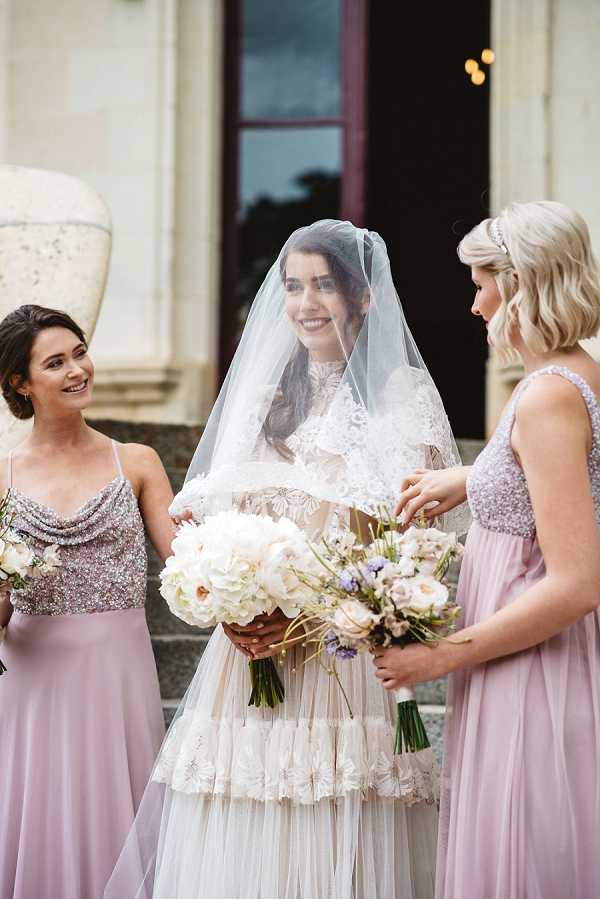 A portrait-style shot of a bride standing between two bridesmaids on the steps of what appears to be a classical stone building with ornate architectural details. The bride wears a tiered lace and tulle gown with a cathedral-length veil draped over her head, and carries a large bouquet of white peonies and hydrangeas. Both bridesmaids wear floor-length mauve dresses with sequined or beaded bodices — one with spaghetti straps and a draped neckline, the other with wider straps — and hold bouquets of blush garden roses, lavender, and mixed soft-toned blooms. The overall styling theme is classic with a vintage-inspired edge, combining intricate lacework, soft mauve tones, and white and blush florals. The bridesmaids are interacting warmly with the bride, and the composition is a medium shot capturing all three figures from approximately the waist up.