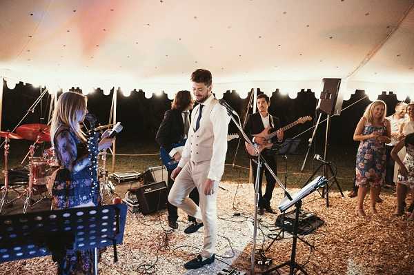 A live band performs during a wedding reception inside a large white sailcloth or marquee tent. The groom, wearing a cream three-piece suit with a dark tie and navy loafers, dances energetically at center stage in front of a microphone stand. The band includes a female vocalist in a floral dress, a guitarist in a dark jacket, and a drummer partially visible at the rear left, with amplifiers and music stands on a wood chip-covered ground floor. Several wedding guests in floral and formal attire watch and socialize along the right edge of the frame, with warm tent lighting and string lights visible along the tent's inner border. Wide shot capturing the full performance setup and guest interaction.