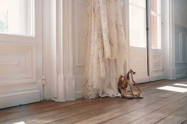 A getting-ready detail shot showing an ivory lace wedding dress hanging in a doorway beside a pair of gold strappy block-heel sandals placed on wide-plank hardwood floors. The dress features intricate floral lace detailing throughout and a flowing skirt. The setting is an indoor room with white wood-paneled walls and doors, likely a chateau or historic venue interior, with natural light streaming through the windows creating soft shadows on the floor. The composition is a wide detail shot at floor level, emphasizing the bridal accessories against the classic white-painted architectural millwork.