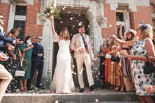A bride and groom exit a brick church or town hall building through a grand stone-framed doorway while approximately 20 guests line the steps on both sides throwing pink and white flower petals. The bride wears a fitted lace white gown and holds a bouquet of yellow and peach flowers raised in the air, while the groom wears a beige/tan suit with a navy tie and dark loafers. Guests are dressed in colorful summer attire including teal, red, and floral-print dresses, suggesting a warm-weather celebration. The scene is captured as a wide mid-shot from a low angle on the steps, conveying movement and energy from the petal toss.