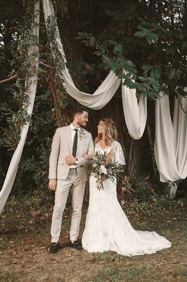 A couple portrait taken outdoors beneath a large tree decorated with draped ivory fabric panels and trailing greenery, creating a boho-style ceremony backdrop. The groom wears a fitted light beige suit with a dark green tie and a small floral boutonniere, while the bride wears a fitted white lace gown with flutter sleeves and a train. The bride holds a loose, organic bouquet featuring dried pampas grass, blush blooms, and olive or eucalyptus greenery in an earthy, neutral palette. The shot is a full-length portrait with both subjects looking at each other and smiling.