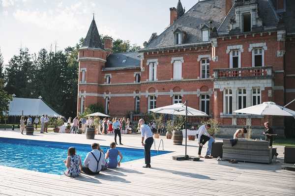 A cocktail hour or outdoor reception taking place on a wooden deck surrounding a large rectangular swimming pool in front of a red brick French chateau with grey slate turrets and white-trimmed windows. Approximately 30-40 guests are spread across the terrace and lawn area, dressed in smart-casual summer attire, with some sitting at the pool's edge and others mingling near white market umbrellas and wooden wine barrel decorations. A white marquee tent is visible to the left, and a lounge seating area with grey wicker furniture is positioned to the right near a bar service station. Wide shot taken from pool level looking toward the chateau facade. Potential venue feature image.