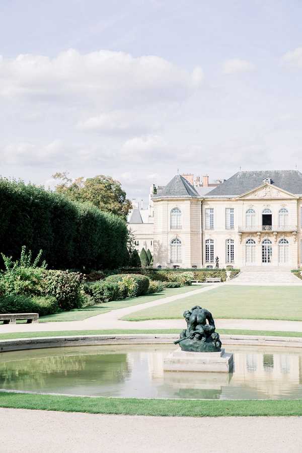 Wide shot of a formal Parisian estate with classical French architecture, featuring cream stone facades, arched windows, and a slate mansard roof. In the foreground, a rectangular ornamental reflecting pool holds a dark bronze sculptural group on a stone plinth. Manicured lawns, clipped hedgerows, topiary cones, and gravel pathways extend between the pool and the building's entrance steps. No people are visible in the frame. Potential venue feature image.