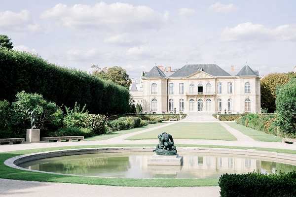 Wide exterior shot of a grand 18th-century Haussmann-style French mansion with a slate mansard roof, cream stone facade, and large arched windows with white shutters. The formal garden in the foreground features a circular reflecting pool with a bronze sculptural group at its center, flanked by manicured lawn, trimmed hedgerows, and topiary. No wedding party or guests are visible in this image. Potential venue feature image.