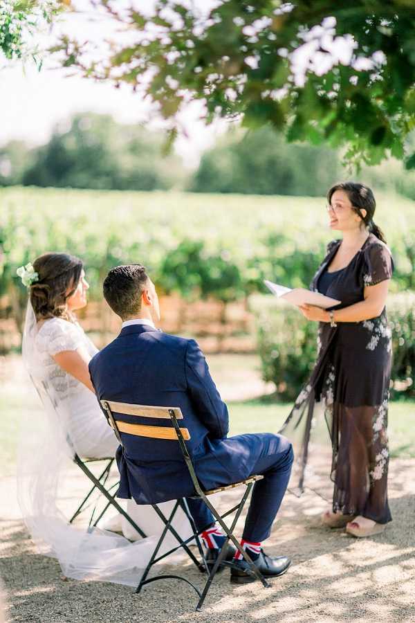 An outdoor wedding ceremony taking place in front of a vineyard, with the couple seated on green metal folding chairs facing an officiant who is reading from papers. The bride wears a lace short-sleeved white gown with a veil and has white flowers in her upswept hair, while the groom wears a navy suit with Union Jack-patterned socks visible above his black shoes. The officiant is dressed in a dark floral maxi dress and glasses. The shot is a medium portrait taken from slightly behind and to the side of the couple, with warm afternoon light filtering through tree branches overhead.