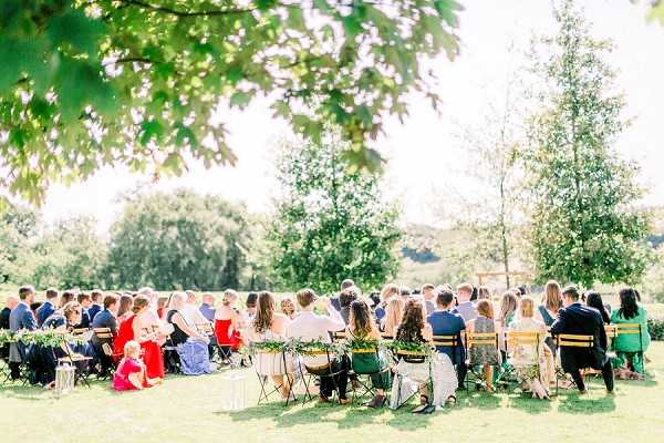 An outdoor wedding ceremony taking place on a grass lawn, with approximately 60–80 guests seated in rows of wooden and folding chairs arranged in an aisle formation. The ceremony appears to be in progress, with guests facing forward toward what appears to be an officiant or couple in the distance. Guests are dressed in a mix of colorful outfits including red, blue, green, and neutral tones. The chair backs are decorated with small greenery arrangements. Wide shot taken from a slight elevation and distance, framed by tree branches in the foreground, giving the scene a garden-party aesthetic in bright natural daylight.