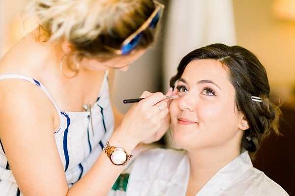 A close-up portrait shot of a bride getting ready indoors, with a makeup artist applying eyeliner or eye makeup using a brush. The bride has dark hair pinned up with a clip and is wearing a white robe, smiling slightly as she looks upward. The makeup artist, a blonde woman wearing a blue and white striped top and a rose gold watch with sunglasses pushed up on her head, leans in to apply the makeup. The setting appears to be a warmly lit indoor room, likely a hotel or getting-ready suite. The image has a bright, airy quality with soft natural lighting.