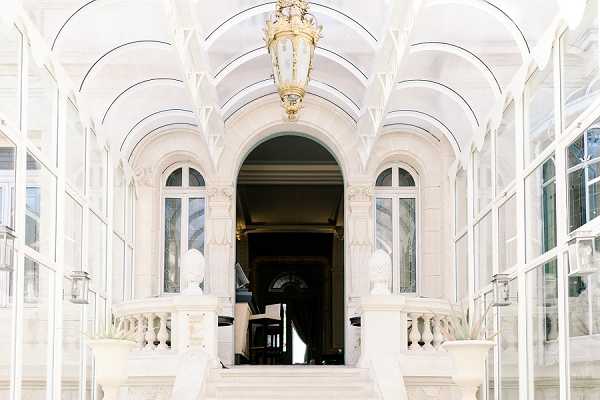 Wide-angle architectural shot of a grand venue entrance featuring a white arched glass-and-iron veranda structure leading to an ornate arched doorway. The covered walkway has a vaulted ribbed glass ceiling and is lit by a large gold lantern pendant light hanging at the center. White marble balustrades flank a set of stairs leading up to the entrance, with classical white sculptural busts positioned on either side of the doorway. No people are visible in the frame. The overall palette is bright white and gold with a light, airy feel from the glass ceiling. Potential venue feature image.