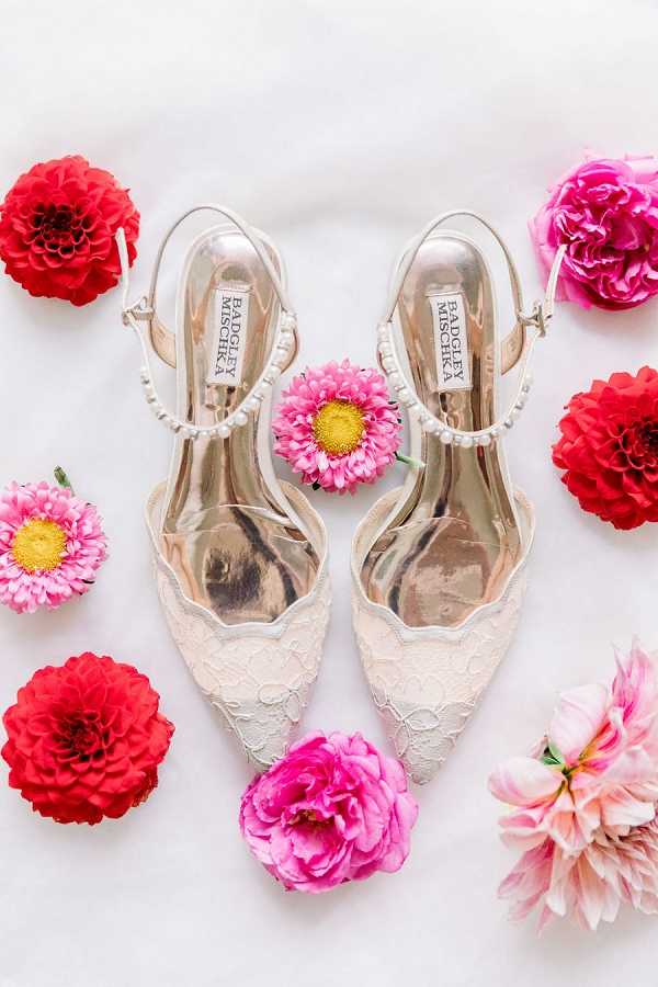 A flat lay detail shot of a pair of Badgley Mischka bridal heels in ivory lace with pearl-embellished ankle straps and gold insoles, arranged on a white surface surrounded by loose flower heads. The flowers include deep red dahlias, hot pink peonies, bright pink gerbera daisies, and light pink dahlias, creating a bold, vibrant color palette against the neutral shoes and white background. The composition is styled with flowers scattered around and between the shoes in a deliberate, decorative arrangement.