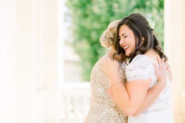 A close-up portrait of two women embracing warmly outdoors, with one laughing joyfully. The bride wears a white lace short-sleeve dress and has a small white or pale green floral hair accessory in her dark hair, which is styled in loose waves. The second woman, likely a bridesmaid or close family member, wears a gold sequined short-sleeve dress with her blonde hair in an updo. The background shows soft-focus white architectural columns and green foliage, suggesting a formal venue terrace or colonnade. The overall color palette is warm and light with a bright, airy feel.