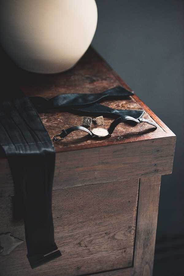 A detail flat-lay style shot of groom's accessories arranged on top of a rustic dark wood chest or trunk, with a large round off-white decorative object partially visible in the upper background. The items include a charcoal grey pleated silk tie draped over the edge, a thin dark leather-strap watch with a round pale face, and two square gold cufflinks. The overall color palette is dark and moody, with charcoal, warm wood tones, and gold accents against a dark grey background. The composition is a close-up detail shot taken at a slight angle, with soft natural light highlighting the textures of the wood and accessories.
