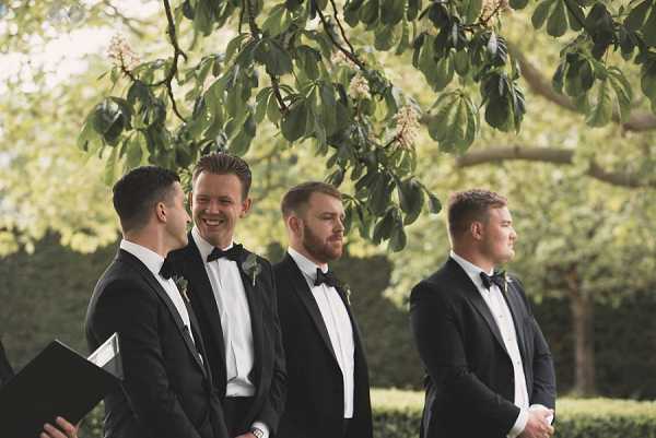 An outdoor wedding ceremony in progress, showing four men standing in a line beneath a leafy tree canopy, with trimmed hedges visible in the background. All four are wearing matching black tuxedos with white dress shirts, black bow ties, and small greenery boutonnieres. Two of the men are laughing and appear to be sharing a light moment, while the others look ahead. One man on the far left is holding a black ceremony folder, suggesting he may be the officiant. The shot is a medium wide portrait capturing the group from approximately the waist up.
